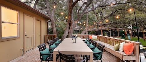 Una terraza al aire libre con una mesa larga y sillas, rodeada de árboles y luces de cuerda.