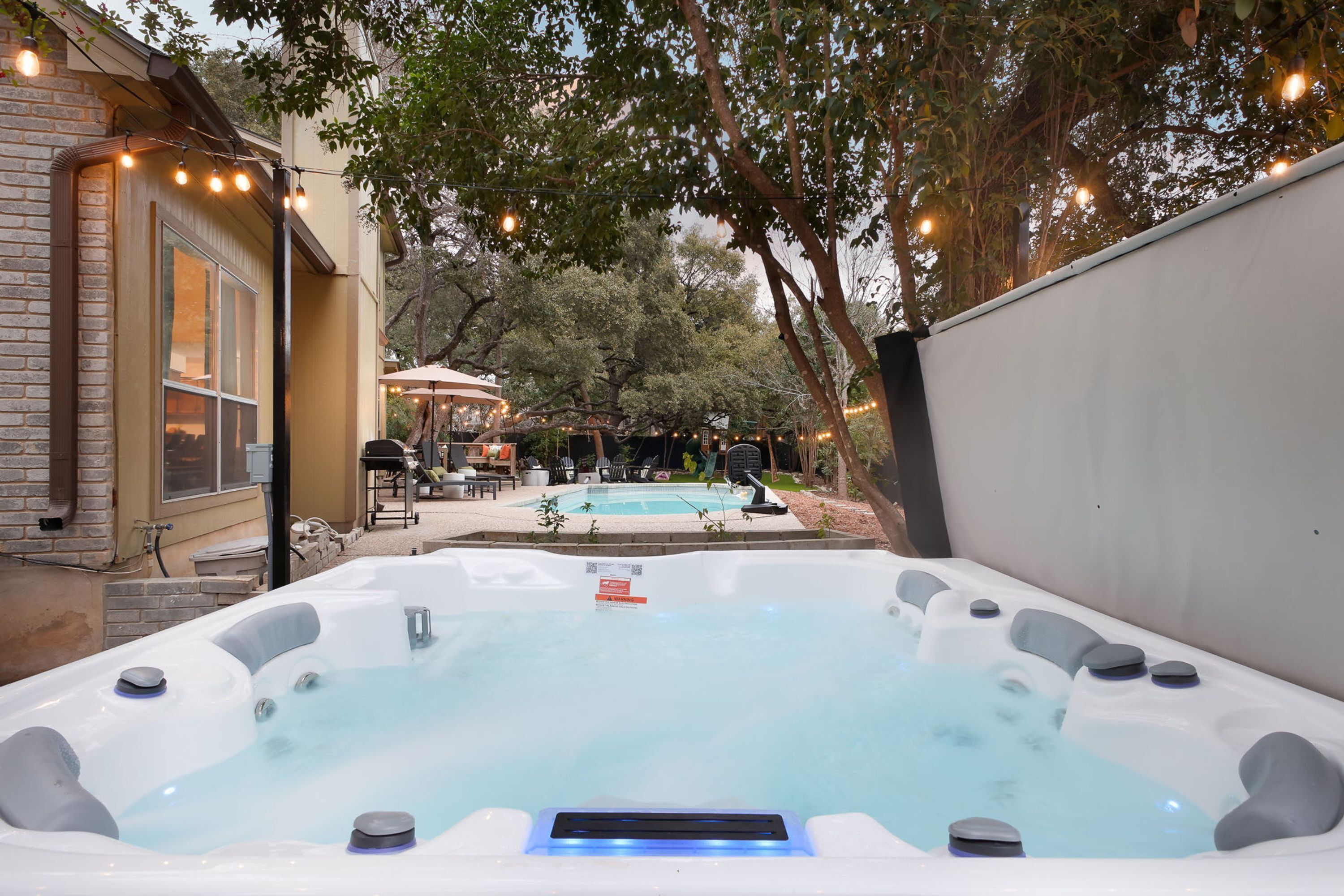 A hot tub with a pool in the background, surrounded by trees and outdoor furniture.