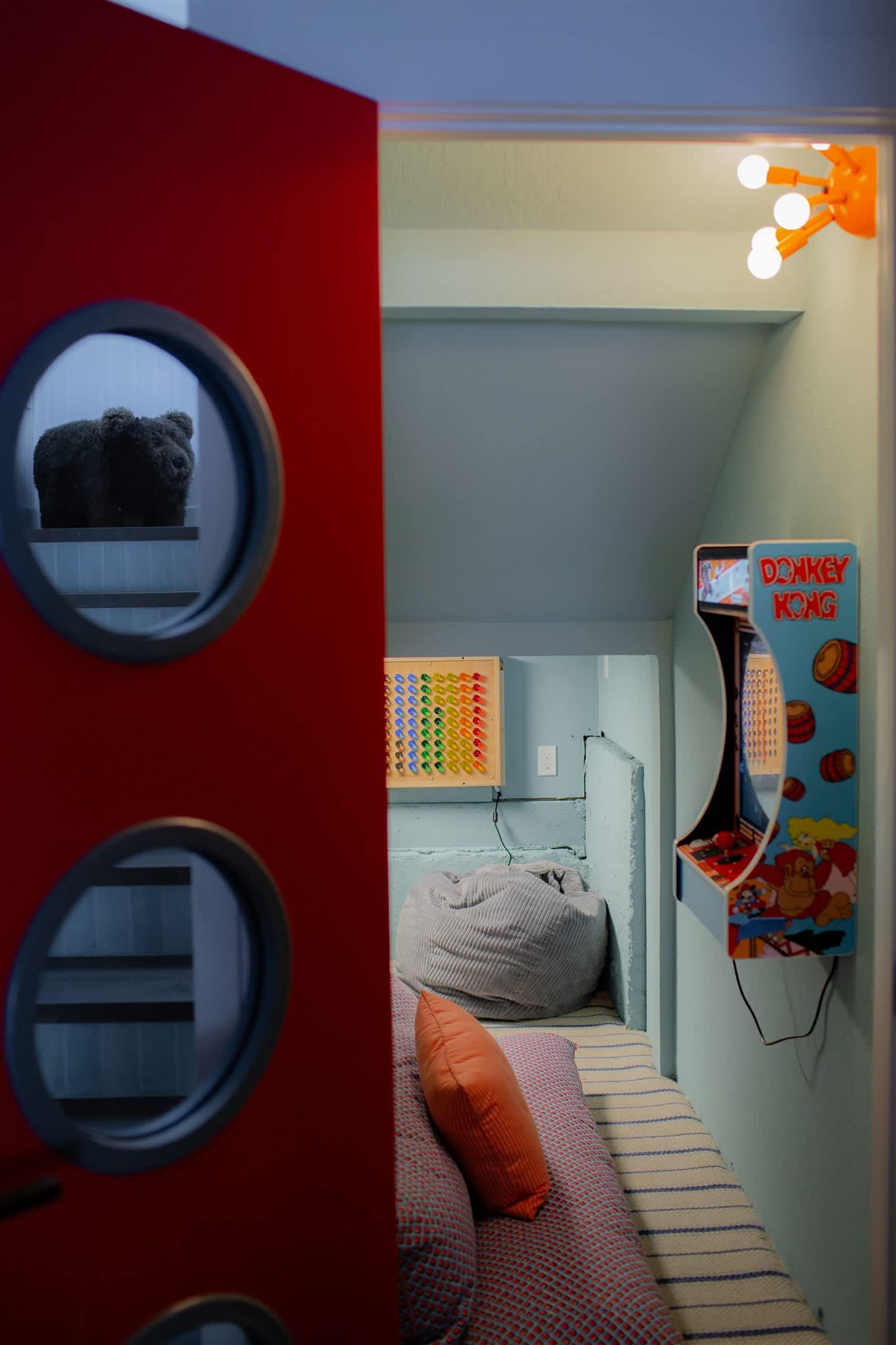 Una habitación con puerta roja, una cama con manta gris y cojín naranja, un escritorio pequeño con una computadora y una máquina de juegos instalada en la pared.