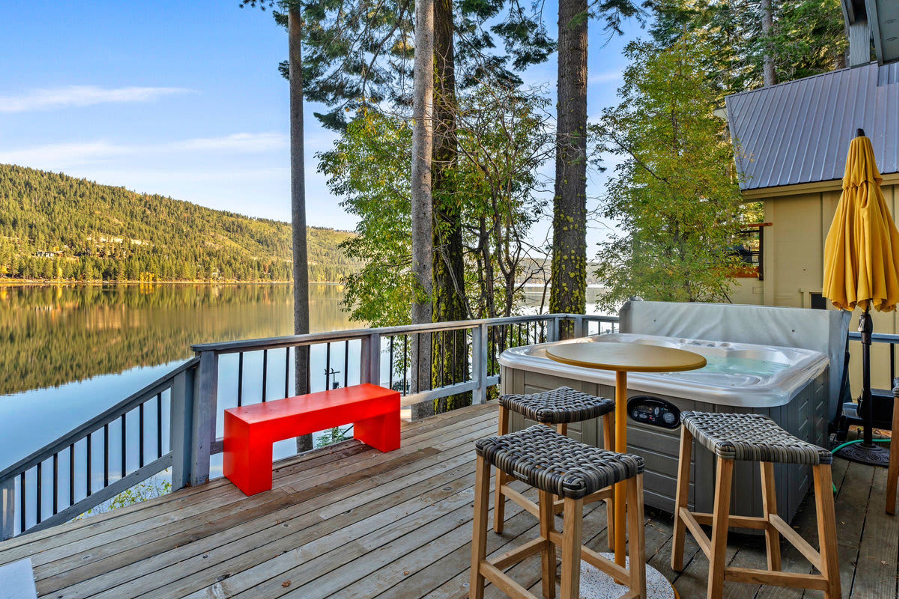 Une terrasse en bois avec un jacuzzi, un banc rouge et des fauteuils en osier donnant sur un lac.
