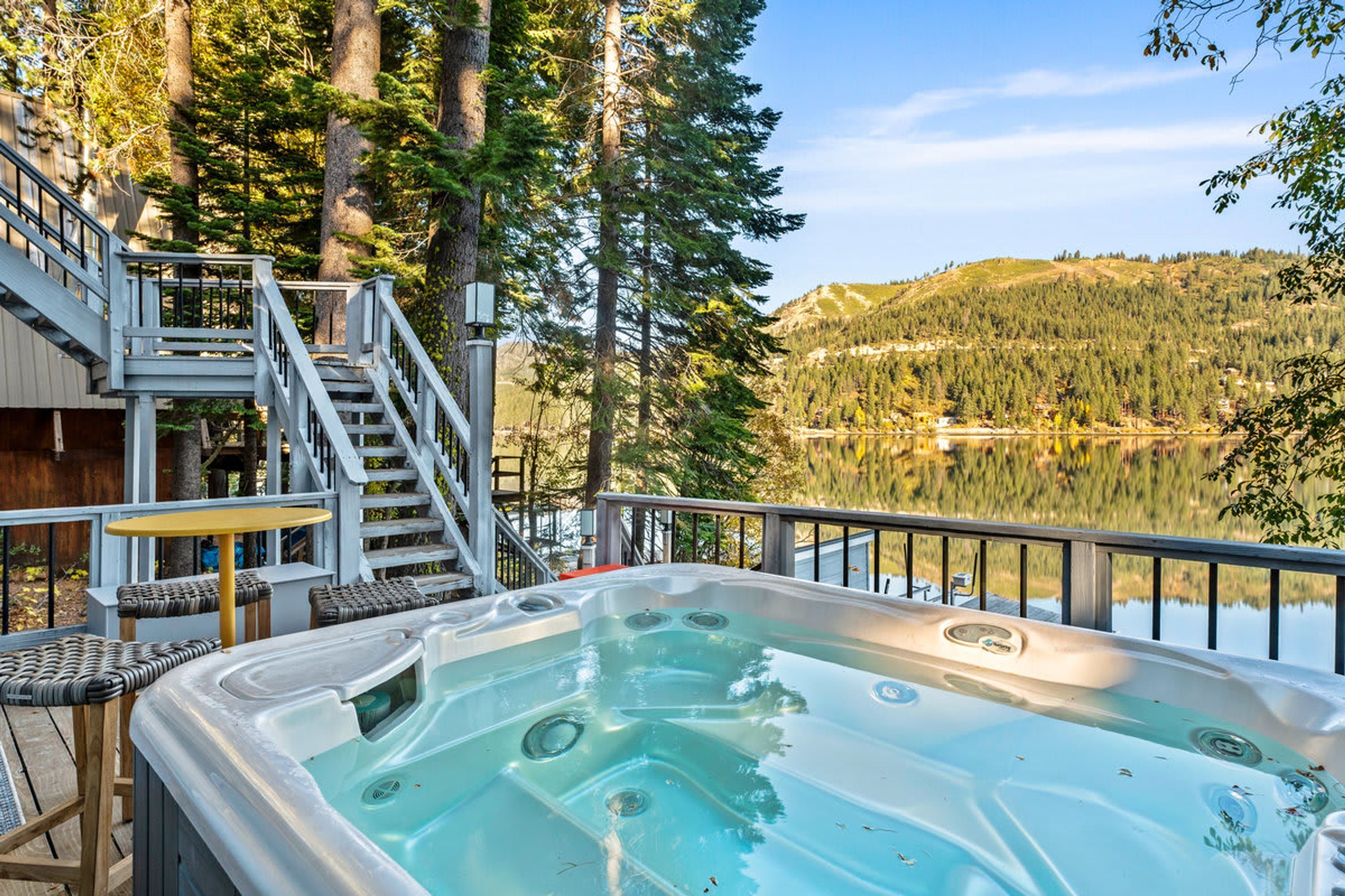 Un jacuzzi con cubierta de madera y sillas, con vista a un lago rodeado de árboles y una montaña al fondo.
