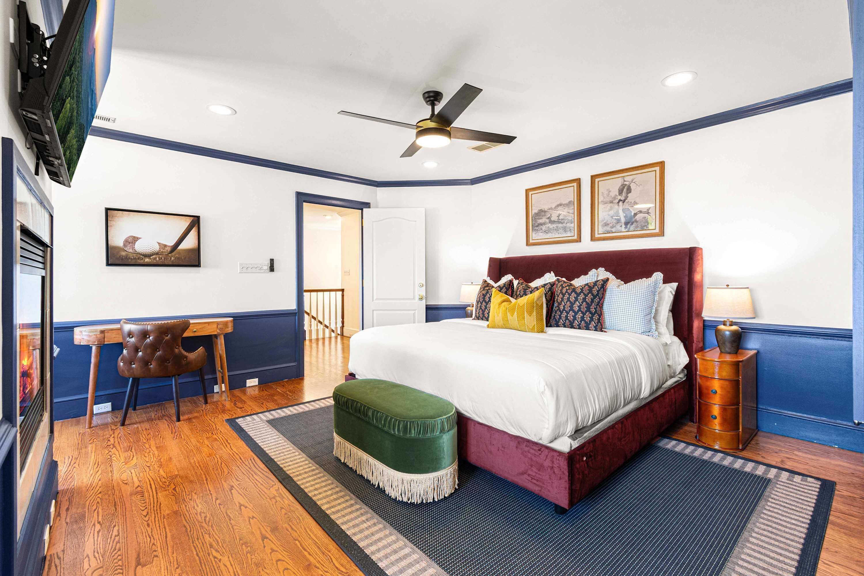 A bedroom with a bed, a desk, a chair, a ceiling fan, and artwork on the walls.