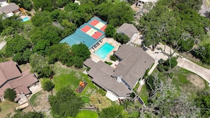An aerial view of a residential area with a swimming pool, tennis courts, and a large house with a sloped roof. - Huge Pickleball Mansion Sleeps 26 Pool Hot Tub (San Antonio)