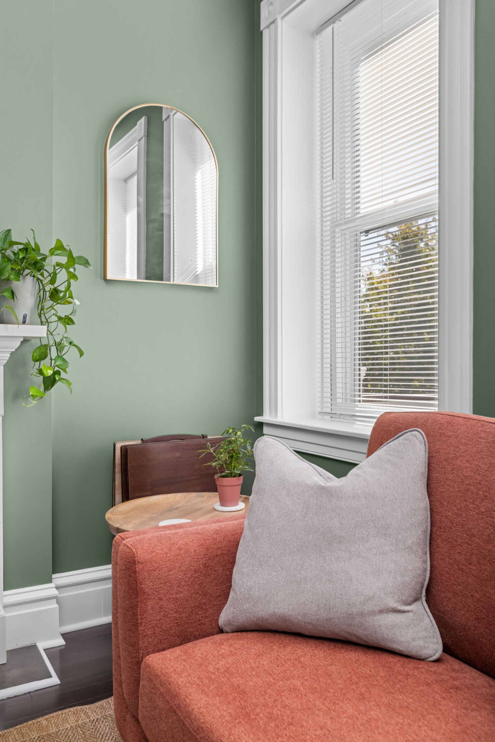 A room with a green wall, a window with blinds, a mirror, a brown chair, a red sofa, and a small table with a plant.