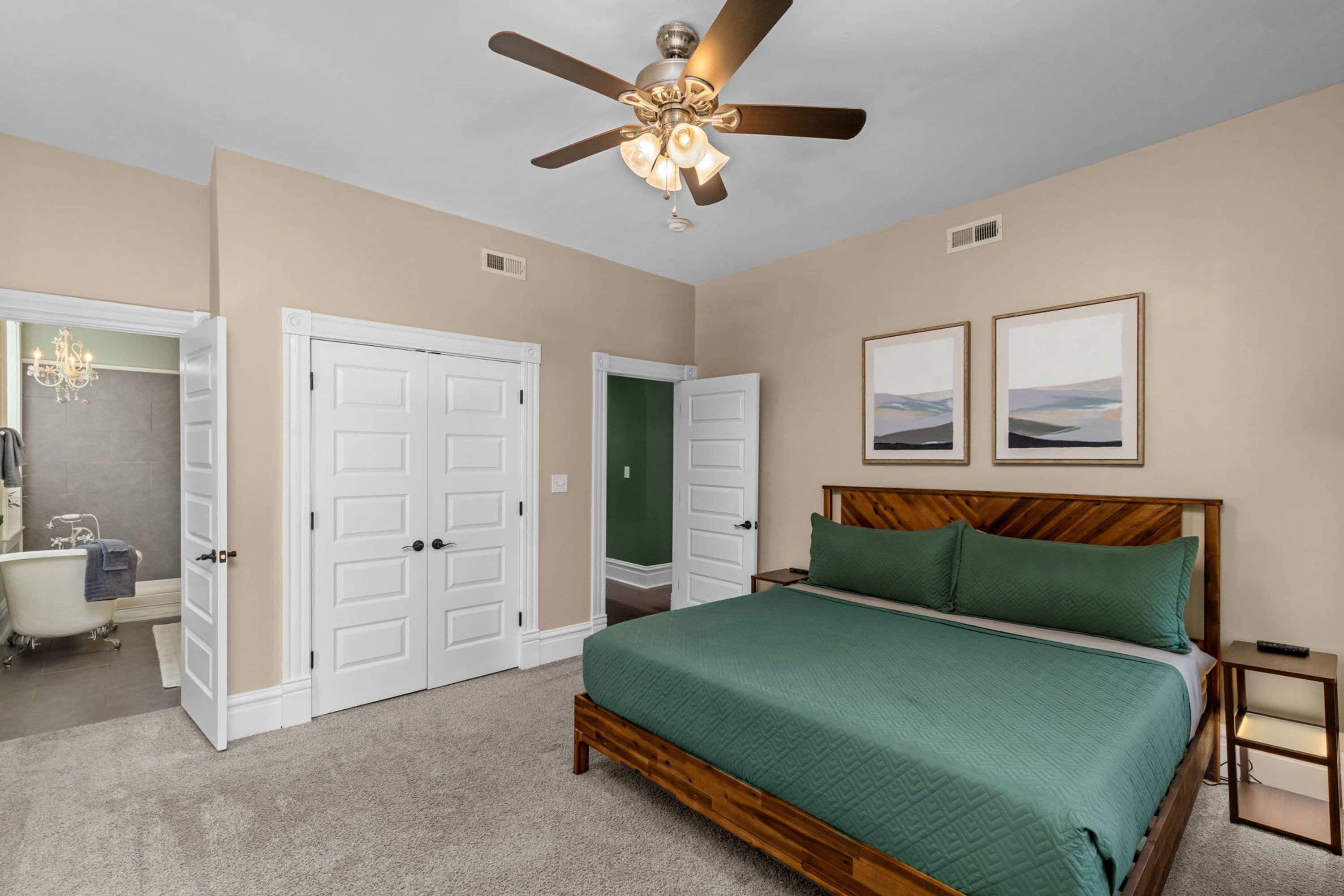A bedroom with a bed, a ceiling fan, two framed pictures, and a bathroom with a bathtub and a chandelier.