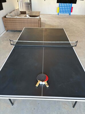 A ping pong table with two paddles in the center, a wicker sofa with cushions, and a blue and red storage unit against a wall. - Bear Butte Luxury Retreat Hot Tub 10 Mins to DT (Sturgis)