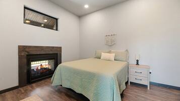 A bedroom with a fireplace, a bed with a light-colored bedspread, a nightstand with a lamp, and a macrame wall hanging.