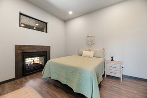 A bedroom with a fireplace, a bed with a light-colored bedspread, a nightstand with a lamp, and a macrame wall hanging. - Bear Butte Luxury Retreat Hot Tub 10 Mins to DT (Sturgis)