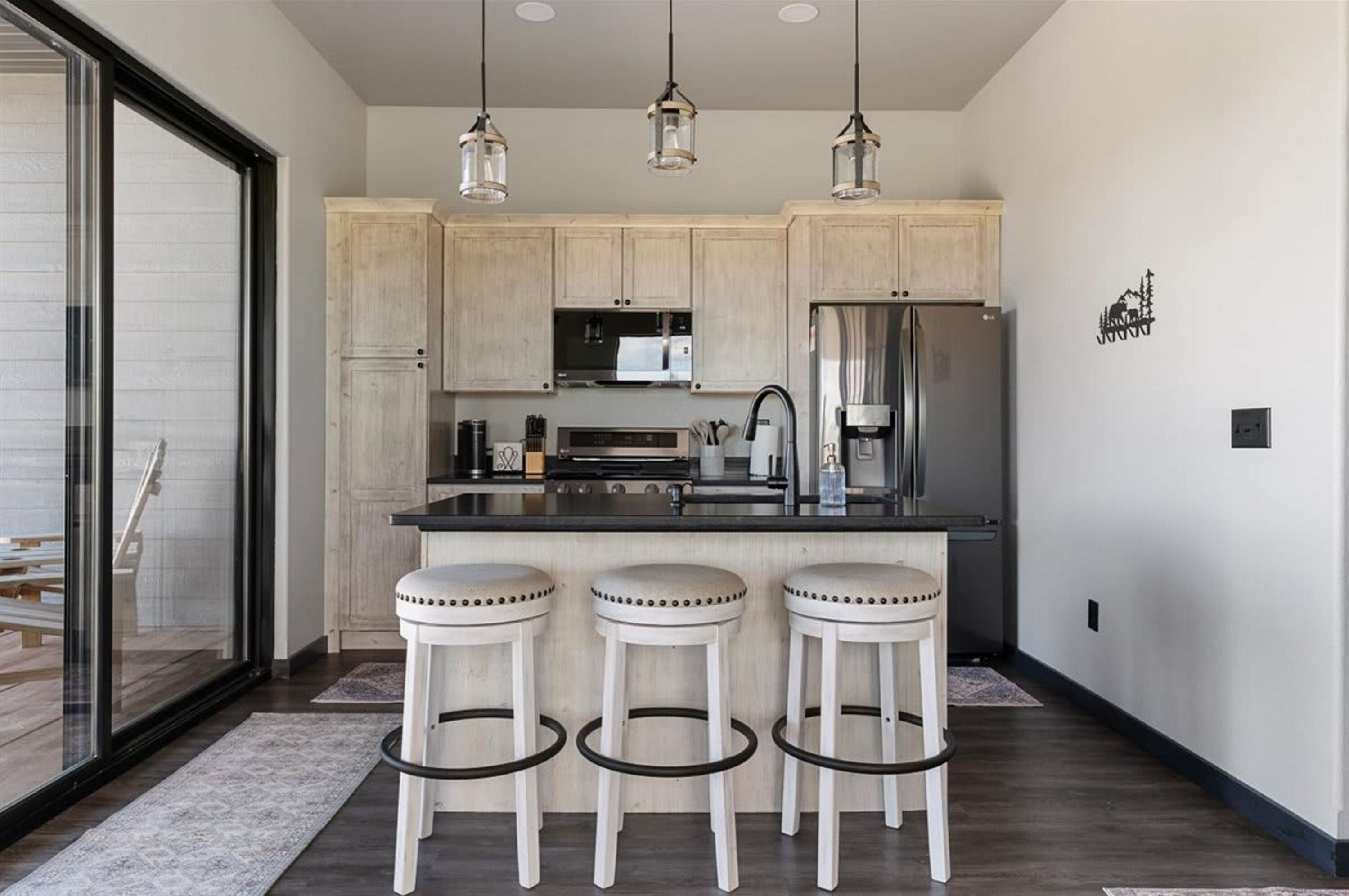 A modern kitchen with wooden cabinets, a stainless steel refrigerator, a microwave, and a coffee maker. There are three white stools with black rings.