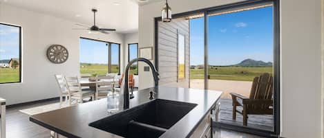 A modern kitchen with a large island, a ceiling fan, and a view of a landscape through a sliding glass door.