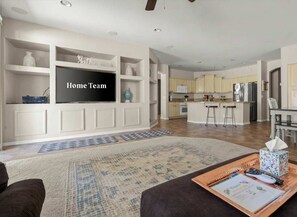 A modern living room with a flat-screen TV, a coffee table with a remote and a tissue box, and a kitchen in the background. - HTD Pool Fire Pit Games Dog Friendly Ping Pong (Chandler)