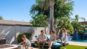 A family by a pool with lounge chairs, a child playing, and a decorative wall. - HTD Pool Fire Pit Games Dog Friendly Ping Pong (Chandler)
