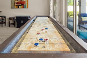 A shuffleboard table with numbered pucks, a painting on the wall, and a view of the outdoors through a window. - HTD Pool Fire Pit Games Dog Friendly Ping Pong (Chandler)