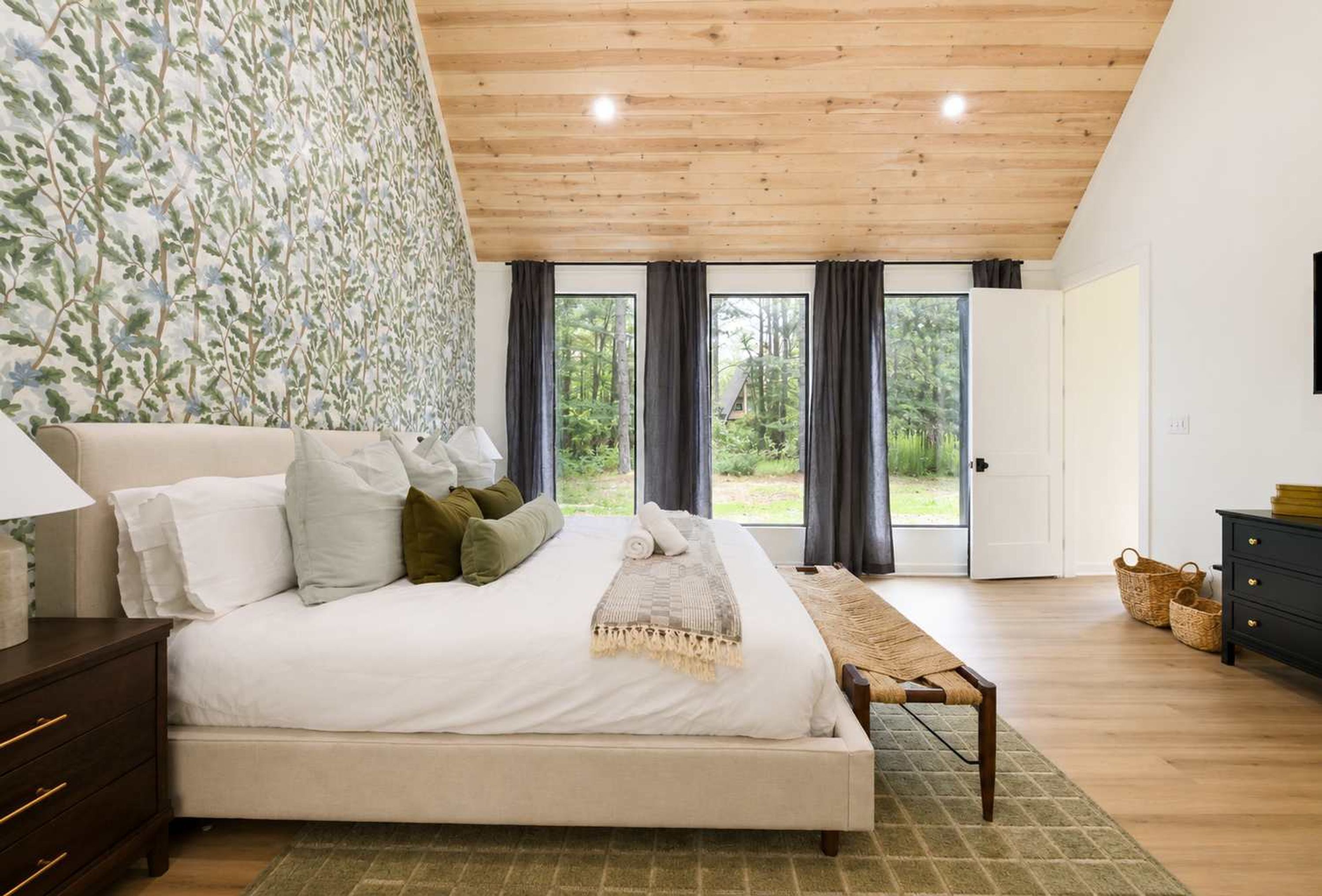 Une chambre à coucher avec un grand lit, une table de chevet, une commode et une vue sur l’extérieur grâce à de grandes fenêtres.