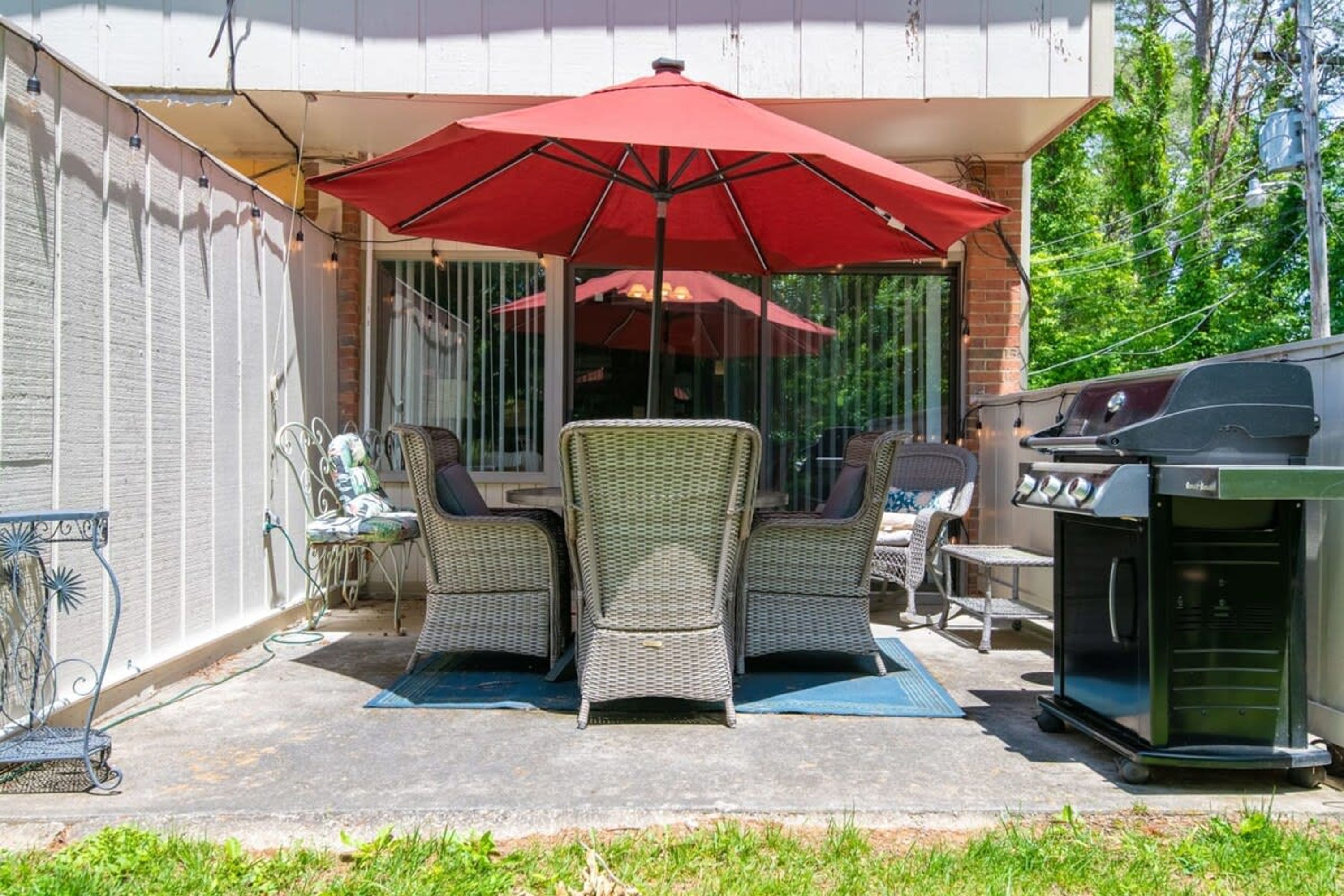 An outdoor patio with wicker chairs, a red umbrella, and a barbecue grill.