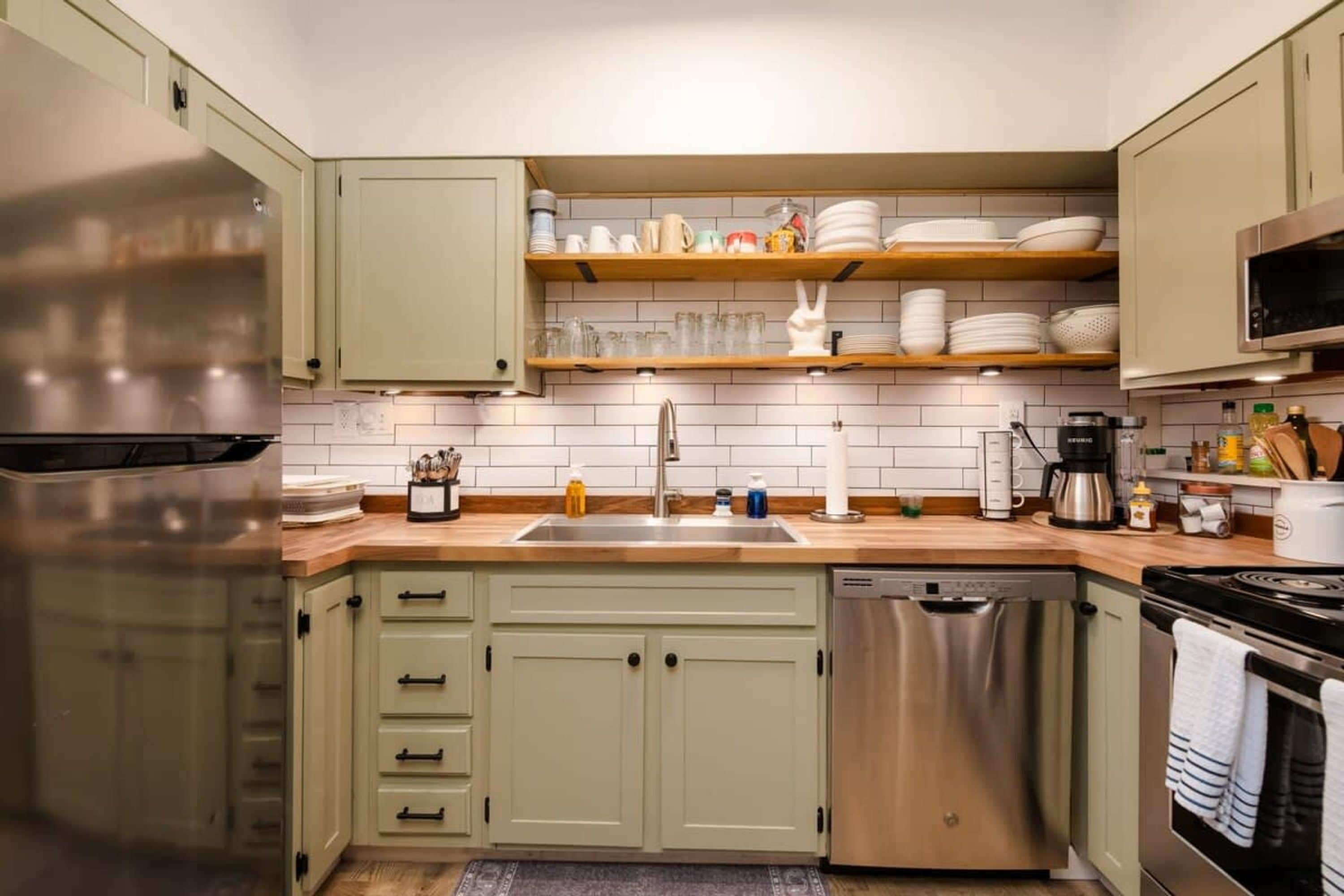 A kitchen with a stainless steel refrigerator, a dishwasher, and a stove. There are wooden shelves above the sink holding dishes and glasses.