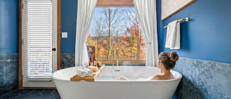 A bathroom with a freestanding bathtub, a window with curtains, and a view of autumn trees.