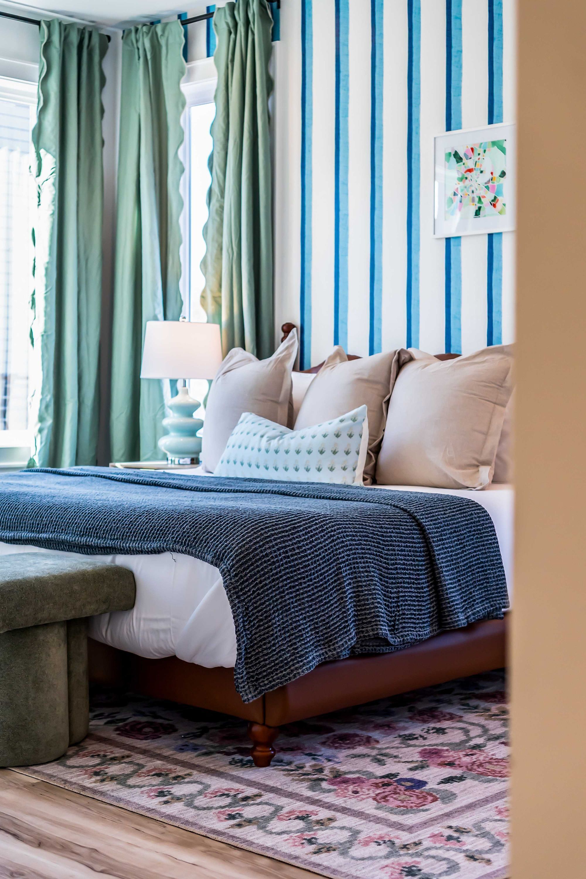 Un dormitorio con cama, almohadas, mesita de noche, lámpara, cortinas y una pared con rayas verticales.