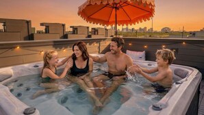 Uma banheira de hidromassagem no terraço, com guarda-sol, emoldurada por um horizonte urbano ao pôr do sol.