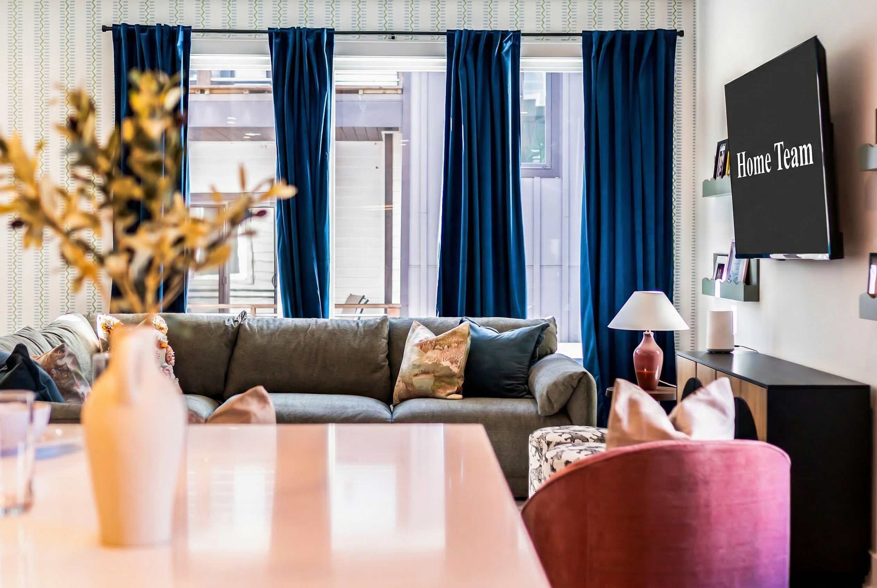 Una sala de estar con un sofá gris, un sillón rojo, un televisor en la pared y una ventana con cortinas azules.