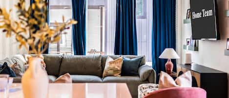 Una sala de estar con un sofá gris, un sillón rojo, un televisor en la pared y una ventana con cortinas azules.