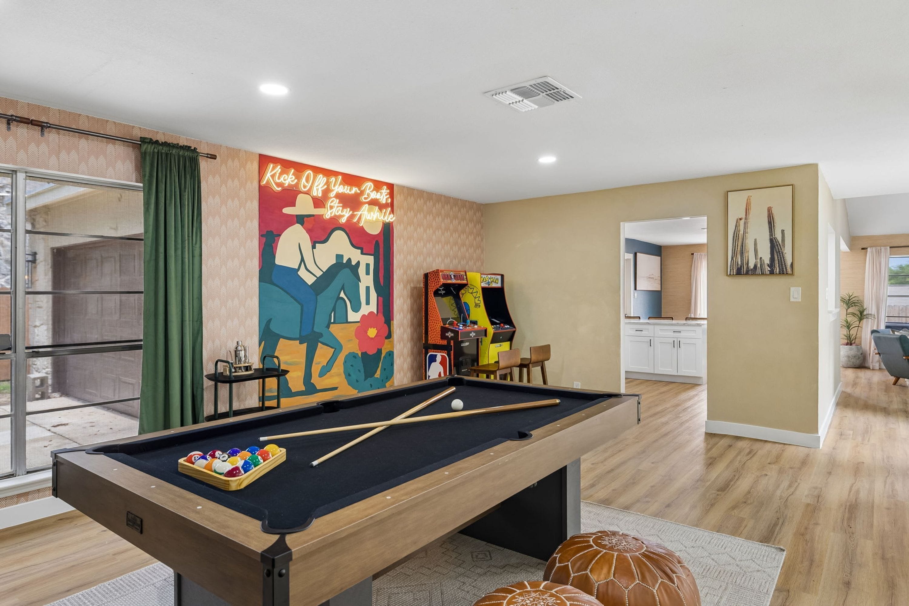 Una mesa de billar con bolas, un mural y una máquina de pinball en una habitación con piso de madera y una cocina al fondo.
