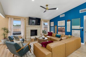 A modern living room with a stone fireplace, a flat-screen TV, and a ceiling fan. - Cowboy Luxe Retreat Pool Putt Putt Pickleball (San Antonio)