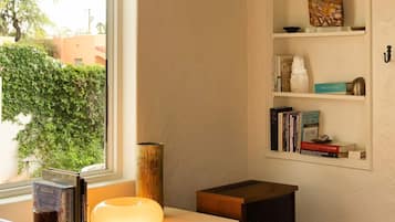 A room with a window, a white desk with a lamp, a bookshelf, and a small black speaker.