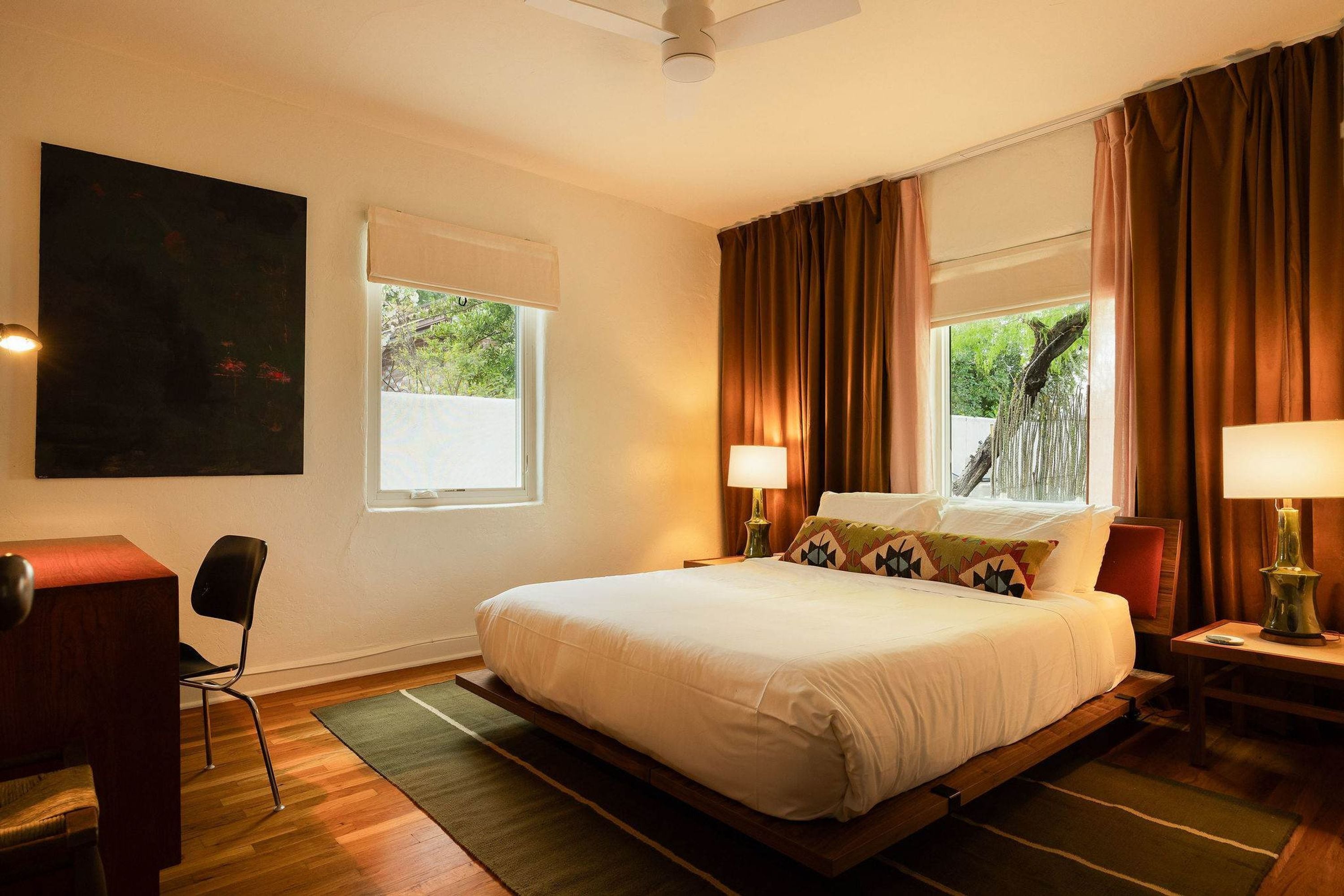 A hotel room with a bed, two bedside lamps, a desk, a chair, and a window with curtains.