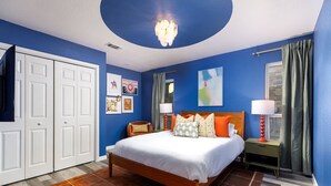 A bedroom with a bed, a nightstand, a lamp, a chair, a wardrobe, and artwork on the walls. - The Sunburst Sleeps 18 Basketball Heated Pool (San Antonio)