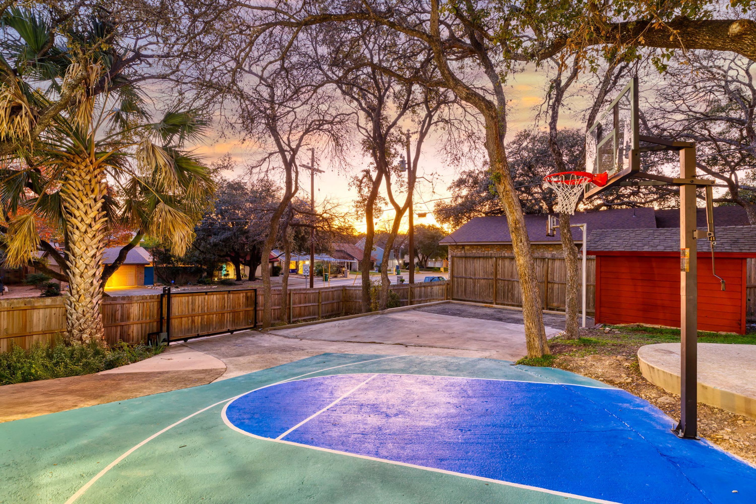 A basketball court with a wooden fence, trees, and a red building in the background.