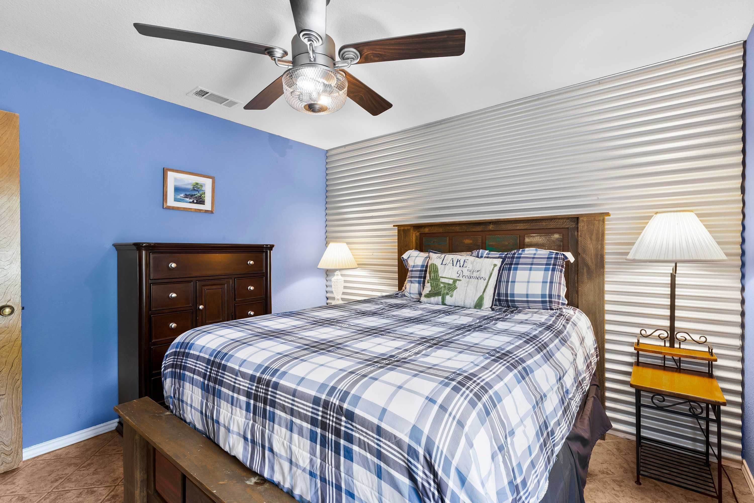 Une chambre à coucher avec un lit, une table de chevet, une lampe, un ventilateur de plafond et un tableau au mur.