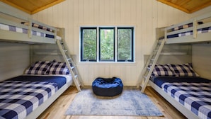 Quarto com duas beliches, janela com vista para árvores, um pufe azul e piso de madeira.
