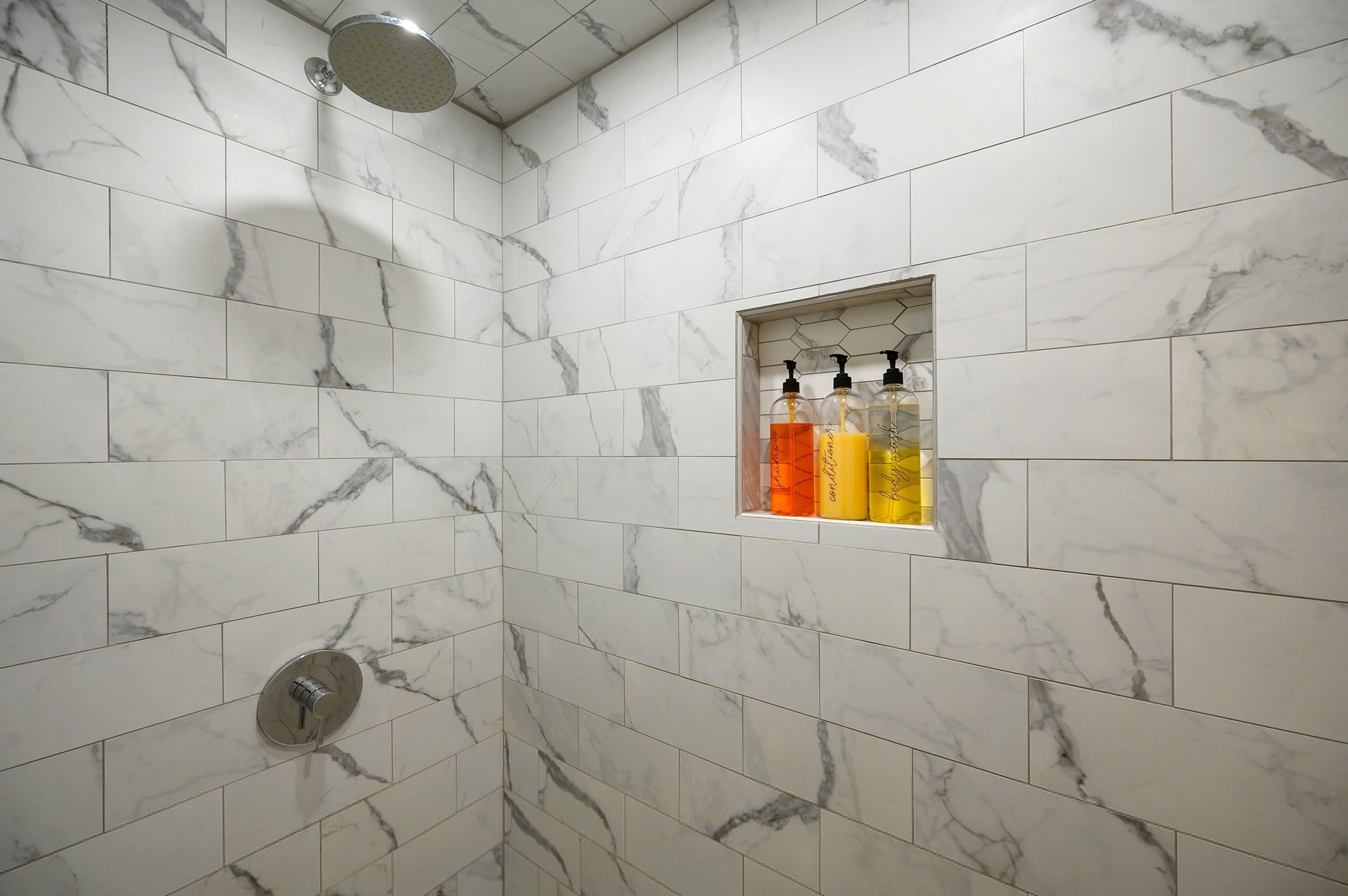 Une salle de bain carrelée de marbre blanc, avec une pomme de douche et une étagère contenant des articles de toilette.