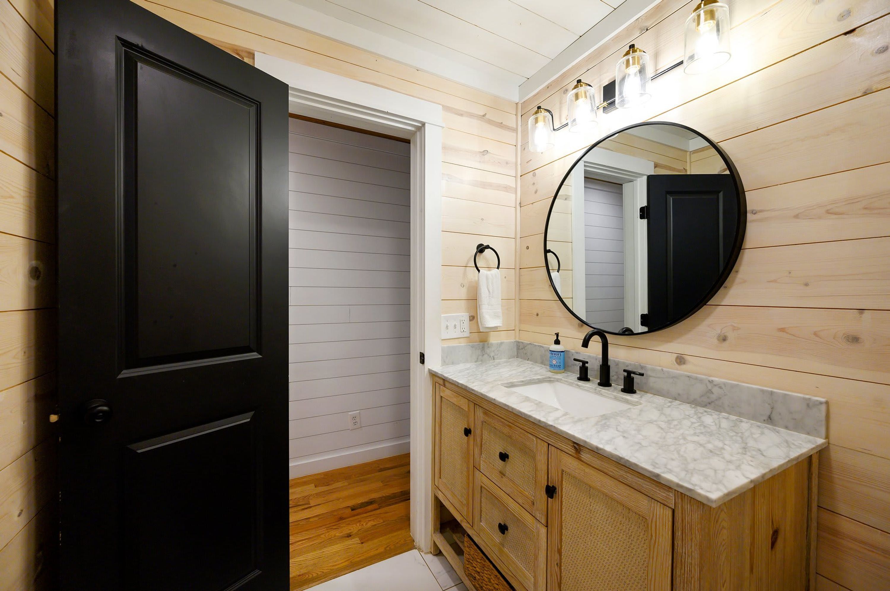 A bathroom with a black door, wooden panel walls, a marble sink, and a round mirror.