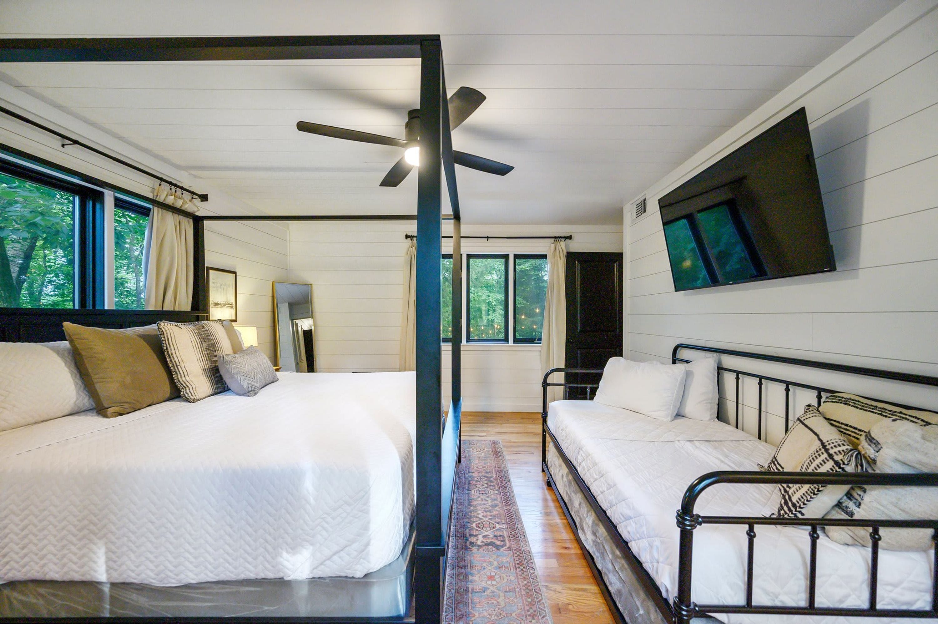 A bedroom with two beds, a ceiling fan, a TV mounted on the wall, and large windows.