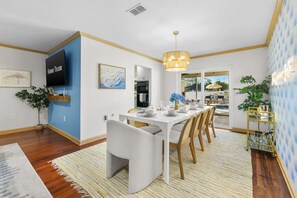 A modern dining area with a white table set for six, a television on the wall, and a view of the outdoors. - Paradise Htd Pool Tiki Bar Hot Tub Games (Seminole)
