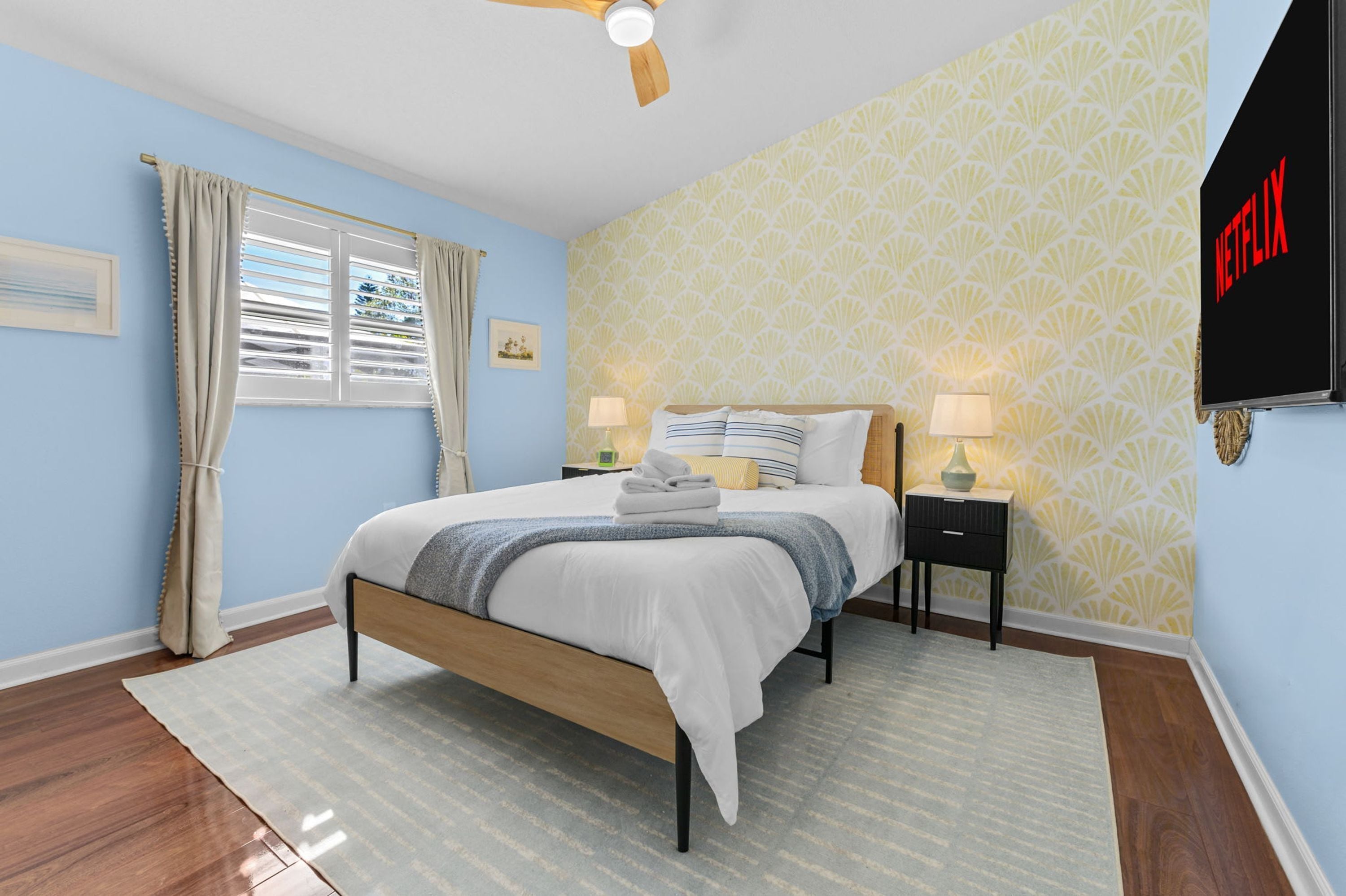 A bedroom with a bed, bedside tables, a ceiling fan, a window with curtains, and a wall-mounted TV.