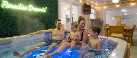 A family in a hot tub with a neon sign reading 'Paradise Dreamin'' in the background.
