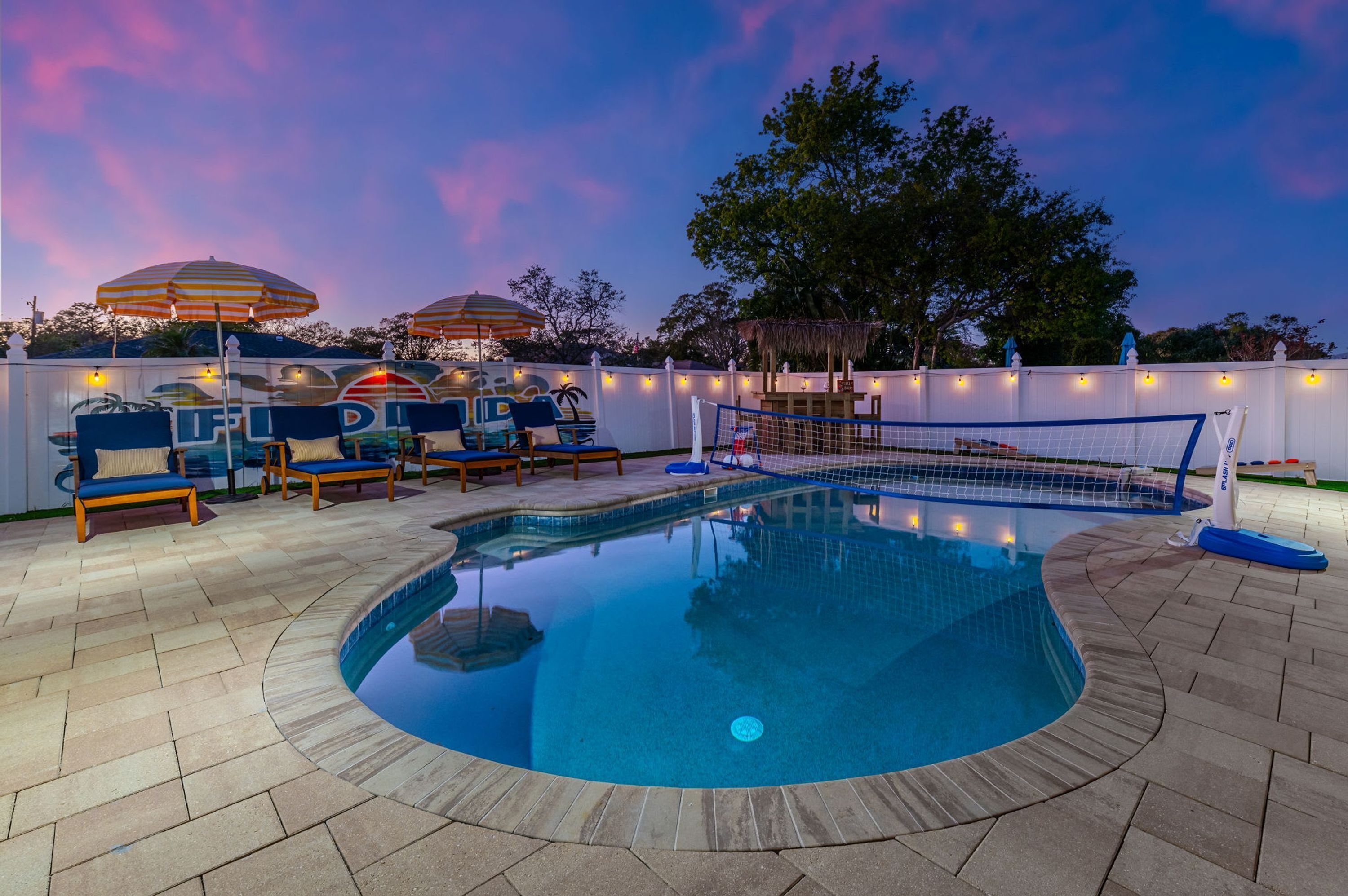 A swimming pool with lounge chairs and umbrellas, surrounded by a fence and string lights.