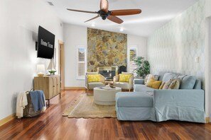 A living room with a ceiling fan, a stone wall, a sofa, a coffee table, and a television. - Paradise Htd Pool Tiki Bar Hot Tub Games (Seminole)