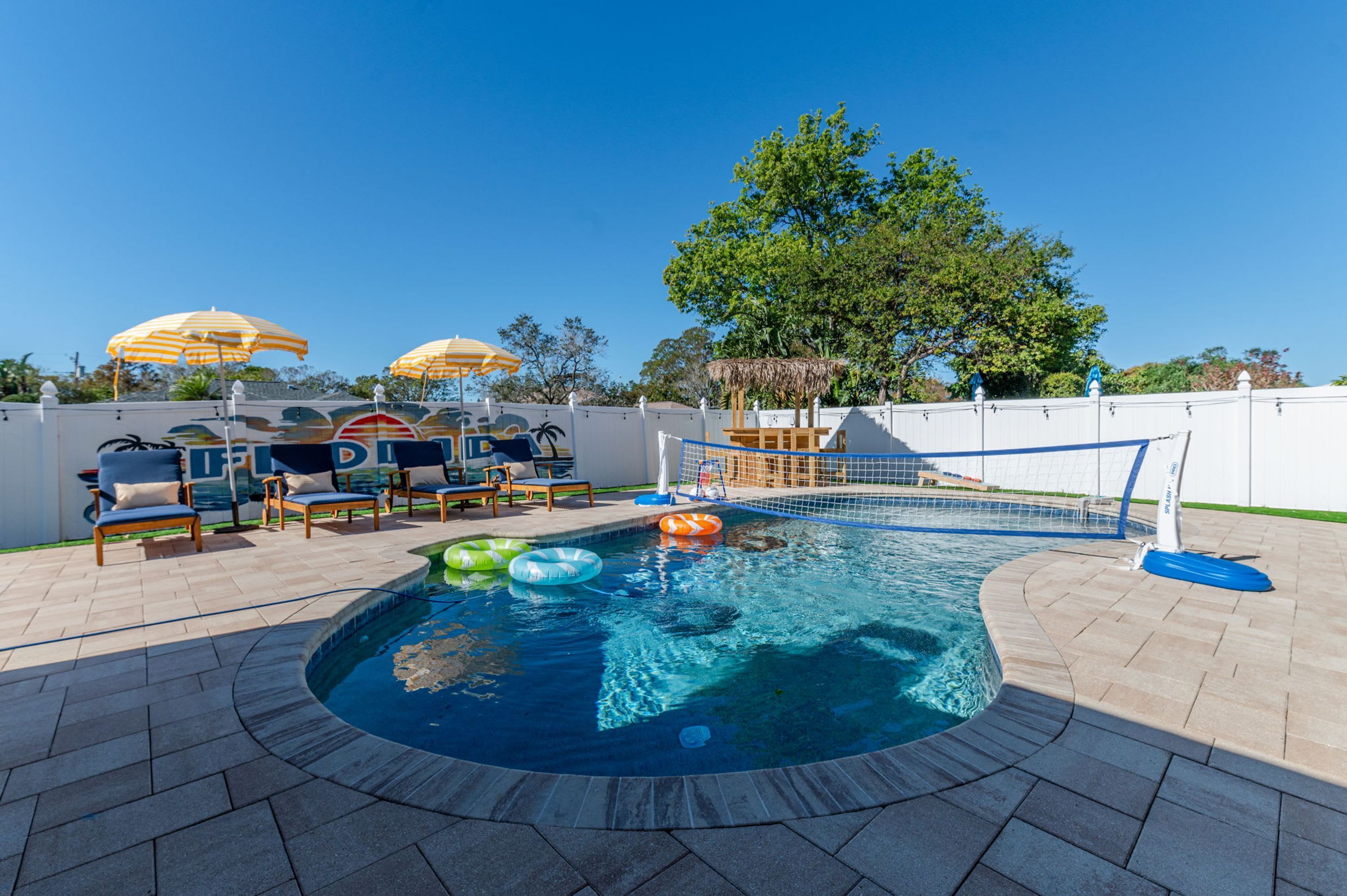 A swimming pool with lounge chairs and umbrellas, surrounded by a white fence and trees.