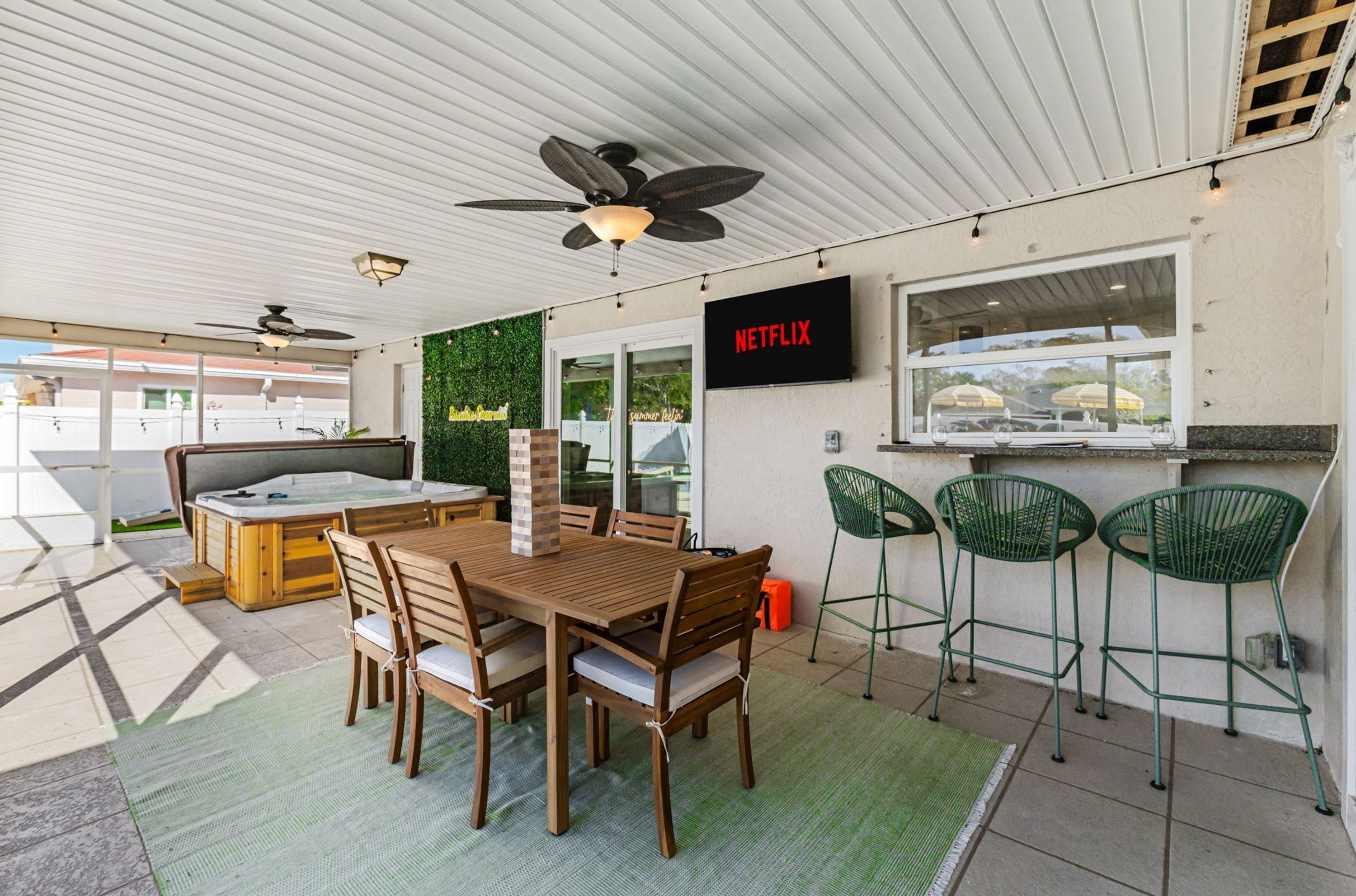 Un espacio exterior techado con una mesa y sillas de madera, un jacuzzi y una pantalla de televisión instalada en la pared que muestra 'NETFLIX'.