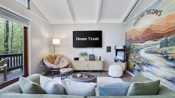 A living room with a large TV screen displaying 'Home Team', a comfortable seating area with a sofa and chairs, a painting on the wall, and a view through a window.