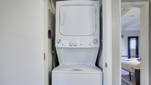 A compact washer-dryer unit installed in a narrow hallway with a bed visible in the adjacent room. - Bluewood Escape Sauna Hot Tub Game Room (Tobyhanna)