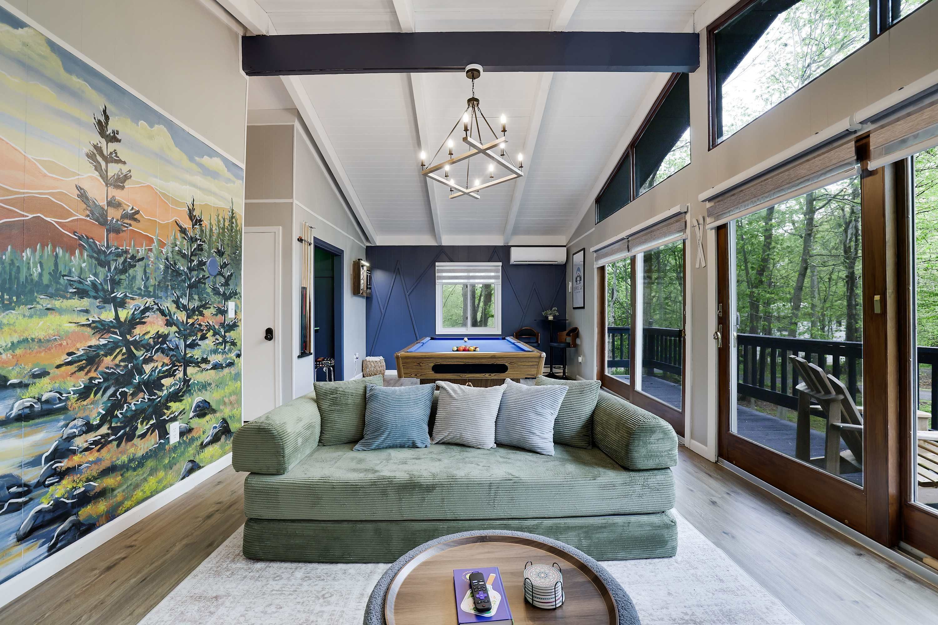 A room with a pool table, a green sofa, a mural, and large windows.