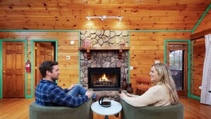 Ein gemütliches Zimmer mit Kamin aus Stein, Holzwänden und zwei zueinander gewandten, grünen Sessel.