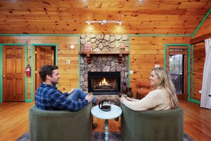 Ein gemütliches Zimmer mit Kamin aus Stein, Holzwänden und zwei zueinander gewandten, grünen Sessel.