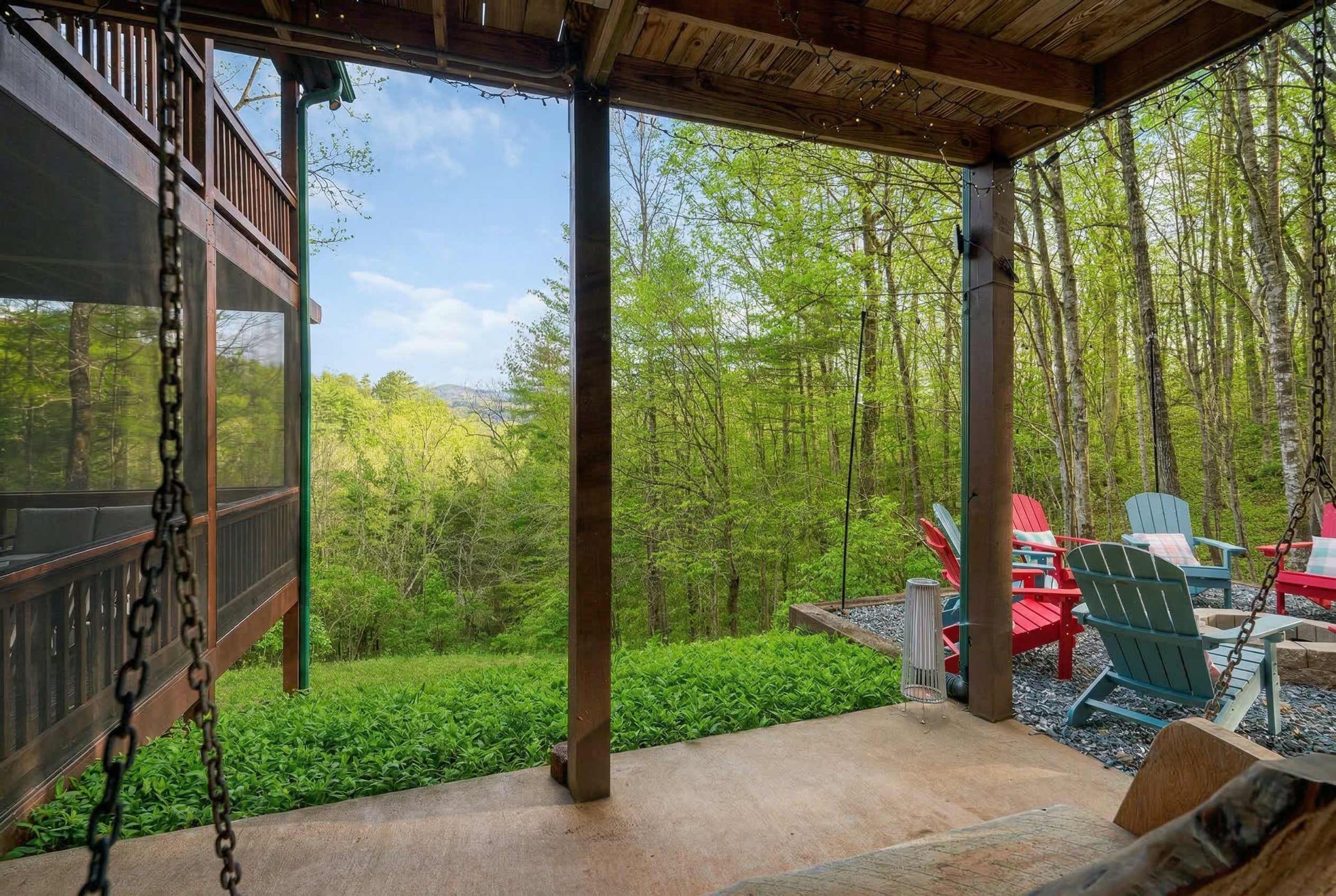 A covered outdoor area with a swing, red and blue Adirondack chairs, and a view of a forested landscape.
