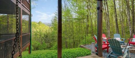 A covered outdoor area with a swing, red and blue Adirondack chairs, and a view of a forested landscape.